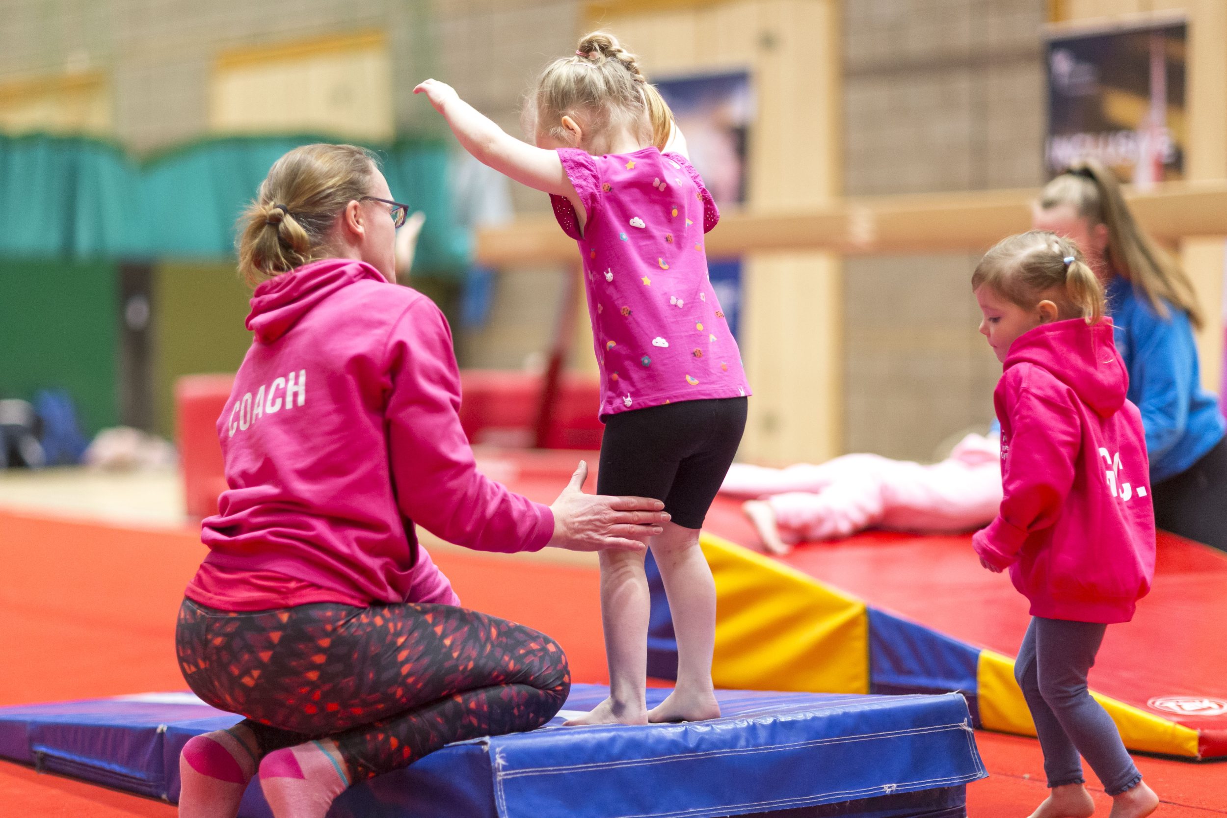 A coach prepares to suport a young gymnast practicing a skill during a pre-school gymnastics class