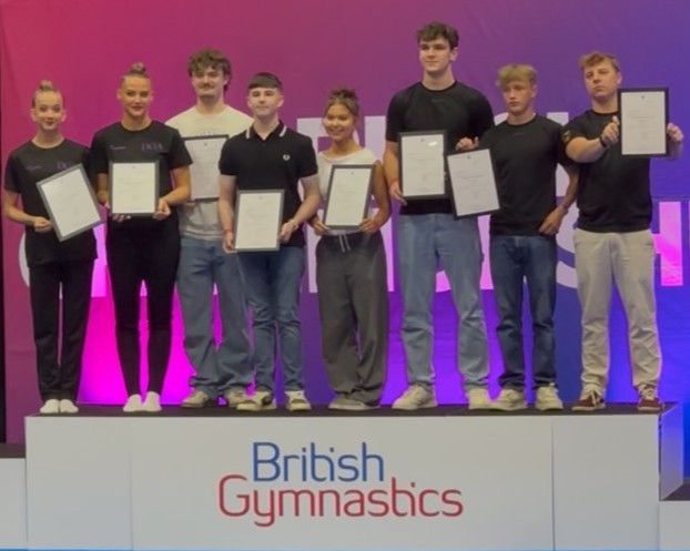 Gemma and Emma stand far left on a podium with six other acrobatic gymnasts holding master gymnast awards